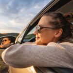 Žena sa naočarima naslonjena na prozor automobila uživa u vožnji kroz prirodu tokom zalaska sunca.
