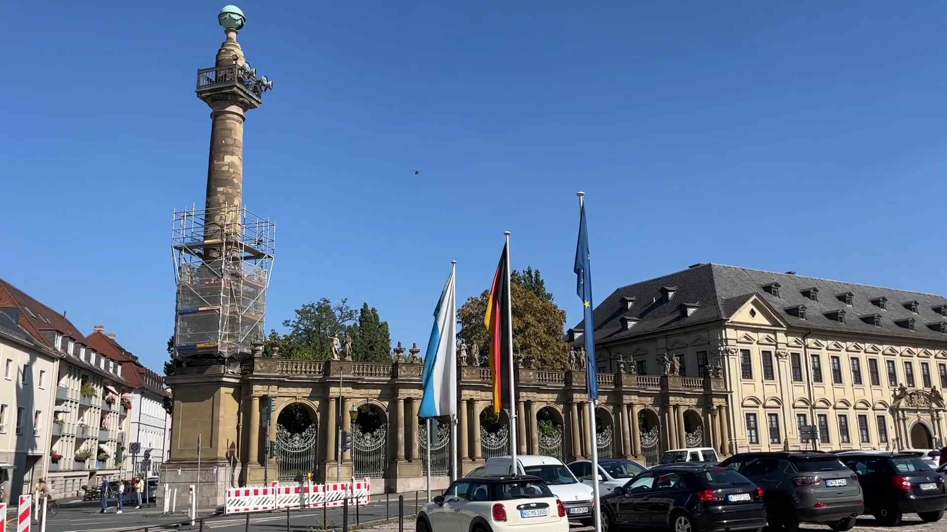 Trg u Würzburgu sa istorijskim stubom, zastavama i baroknim zgradama u pozadini