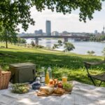 Piknik na zelenoj površini pored reke sa ćebetom, hranom, ručnim frižiderom i pogledom na Beograd